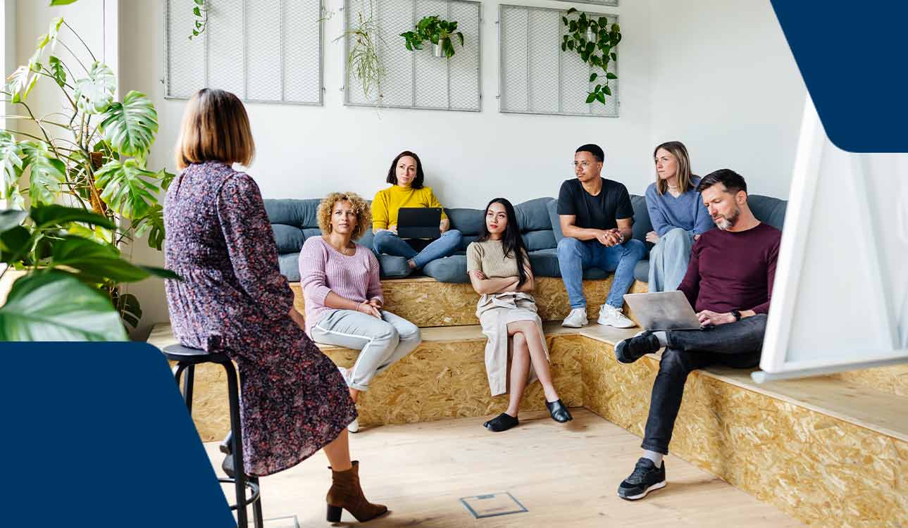 Zu sehen ist ein Team in einem Gespräch, eine Frau sitzt vorne auf einem Hocker und die anderen sechs Team-Mitglieder sitzen vor ihr.