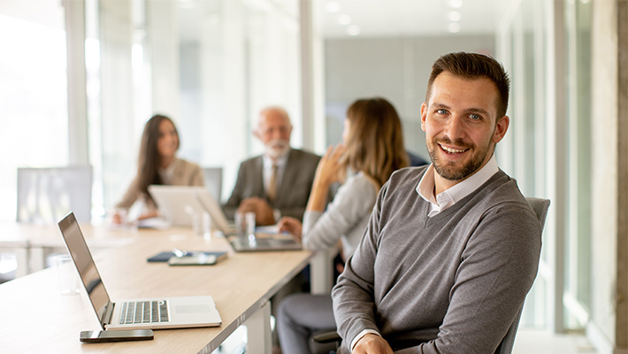 Als Projektteam erarbeitet man seine Projekte gemeinsam, Projektleiter sitzt am Tisch mit Laptop und schaut in die Kamera.