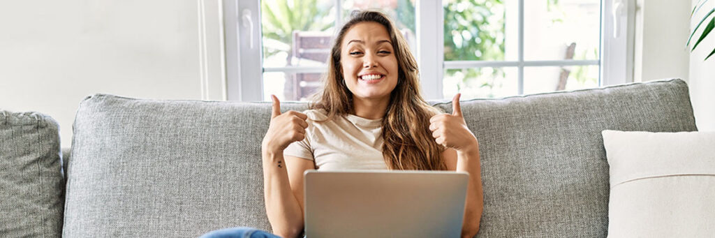 Frau auf Sofa mit Laptop, lächeln, streckt beide Daumen hoch. Symbolhaft für Freude über die Top 5 IT-Berufe.